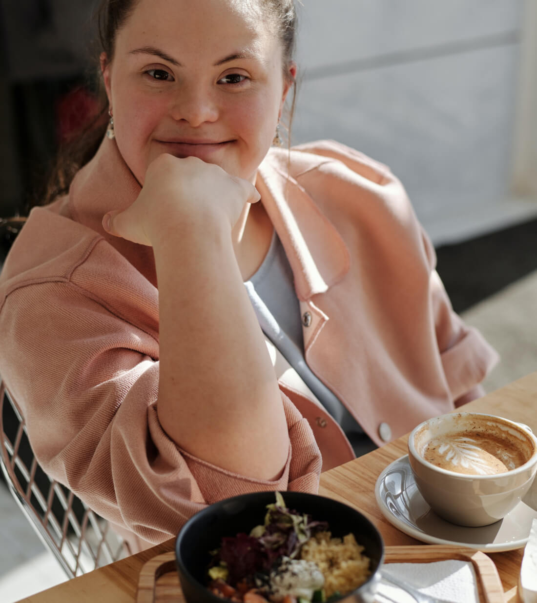 A young woman with dark hair and a light pink jacket smiles, chin resting on her hand, with food and coffee on a table.