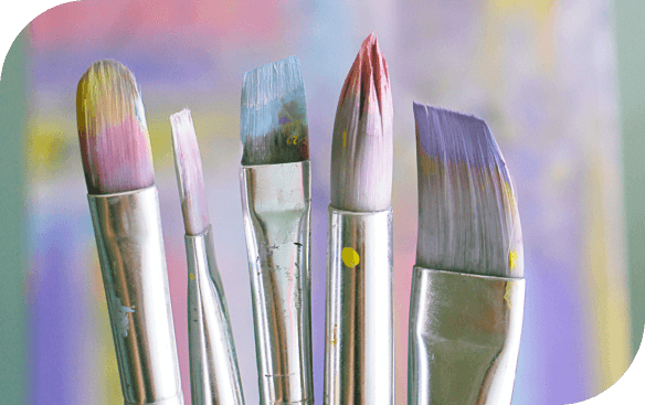 Five paintbrushes with colorful paint on their bristles stand upright against a blurred pastel background.