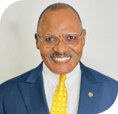 A smiling older Black man with a mustache and glasses wears a blue suit and yellow polka dot tie.