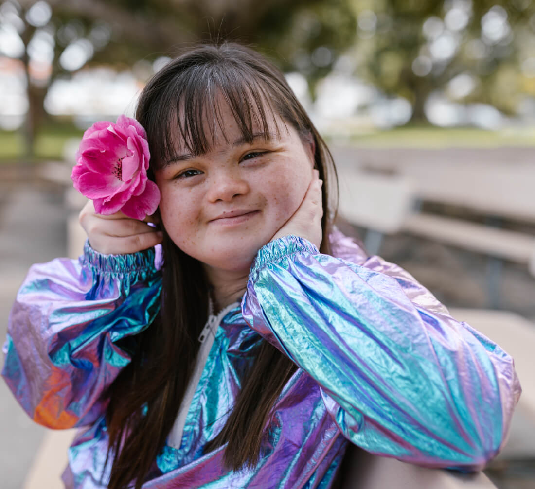 A young woman with dark hair and bangs smiles, holding a pink flower to her ear, wearing a shiny iridescent jacket.