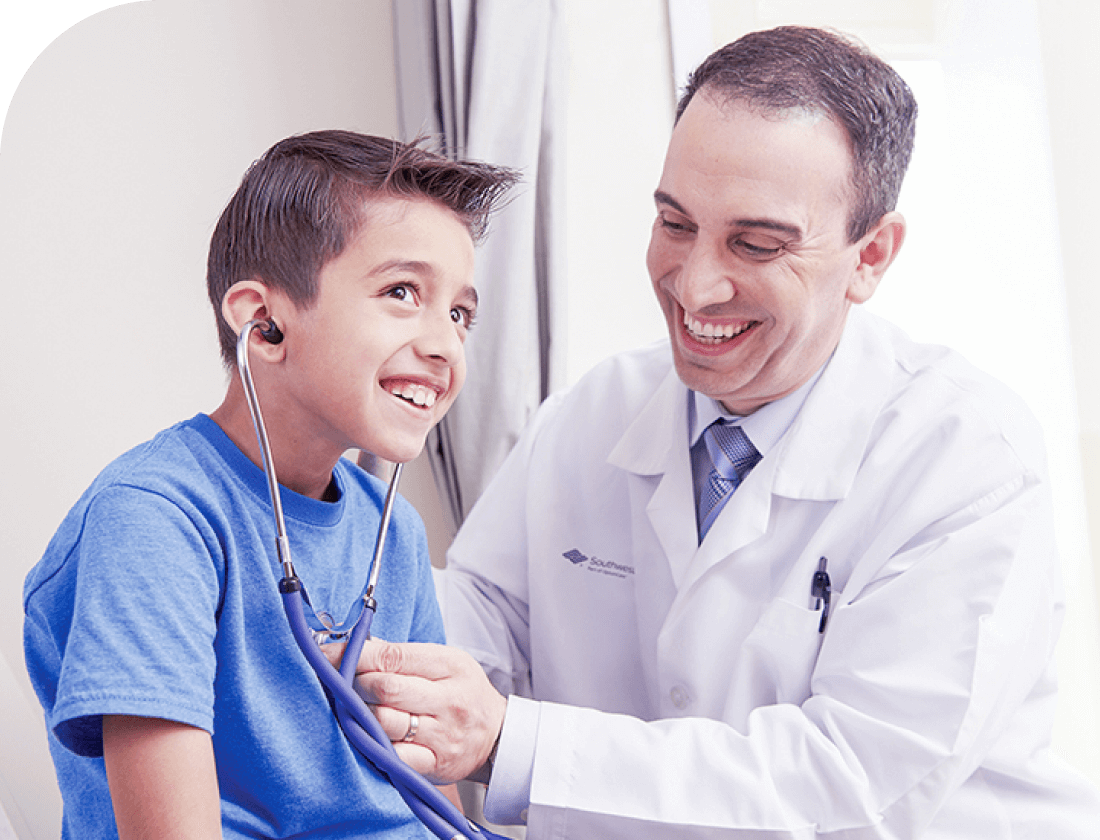 A smiling boy in a blue shirt holds a stethoscope to his chest while a laughing doctor watches.