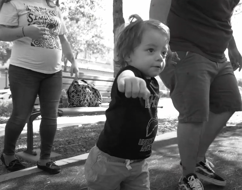 A toddler in a "UNITED STATES" shirt walks forward, with two adults behind them in a park.