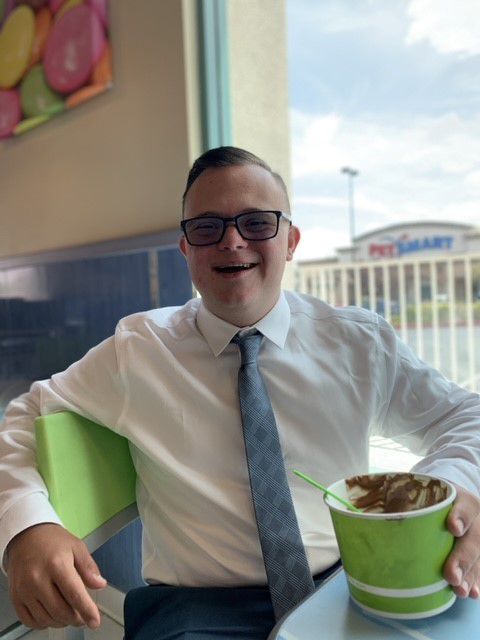 A smiling man in glasses, a white shirt, and a gray tie holds a green cup of chocolate ice cream.