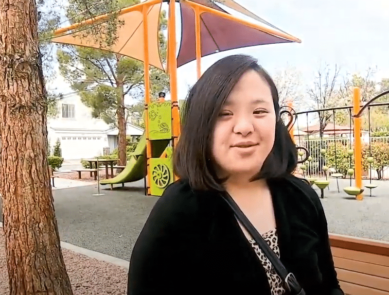 A young woman with dark hair and a black top smiles at the camera in a sunny park with a playground.
