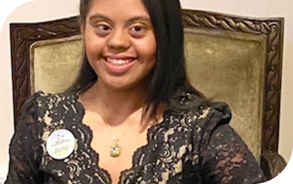 A smiling woman with dark hair and a black lace dress sits in a gold chair, wearing a pin that says "Sophia."