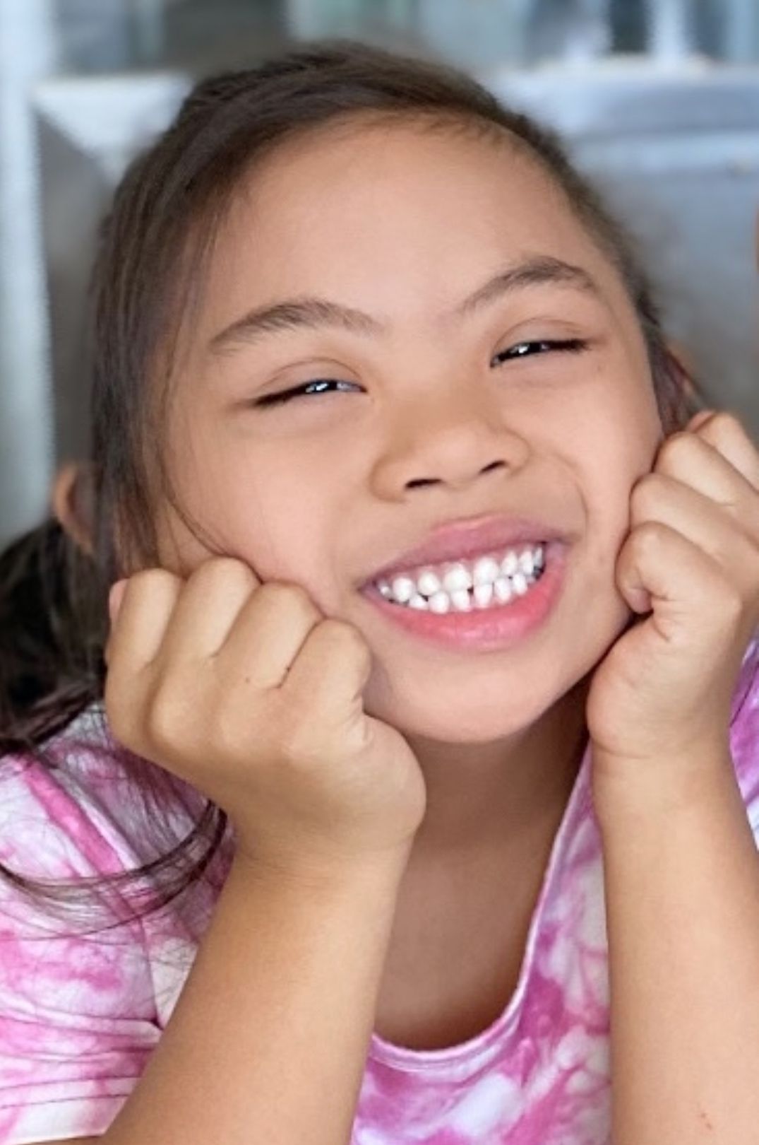 A smiling young girl with dark hair and a pink and white tie-dye shirt holds her hands to her cheeks.