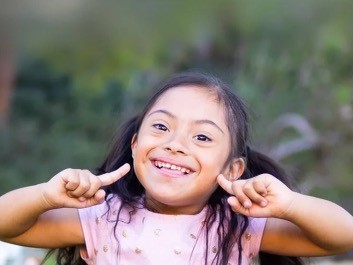 A smiling young girl with long dark hair, wearing a pink top, points to her cheeks with both index fingers.