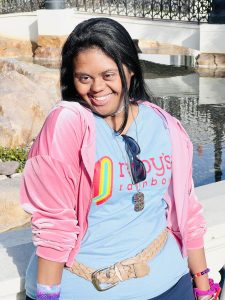 A smiling woman with dark hair wears a pink jacket over a blue "Ruby's Rainbow" t-shirt.