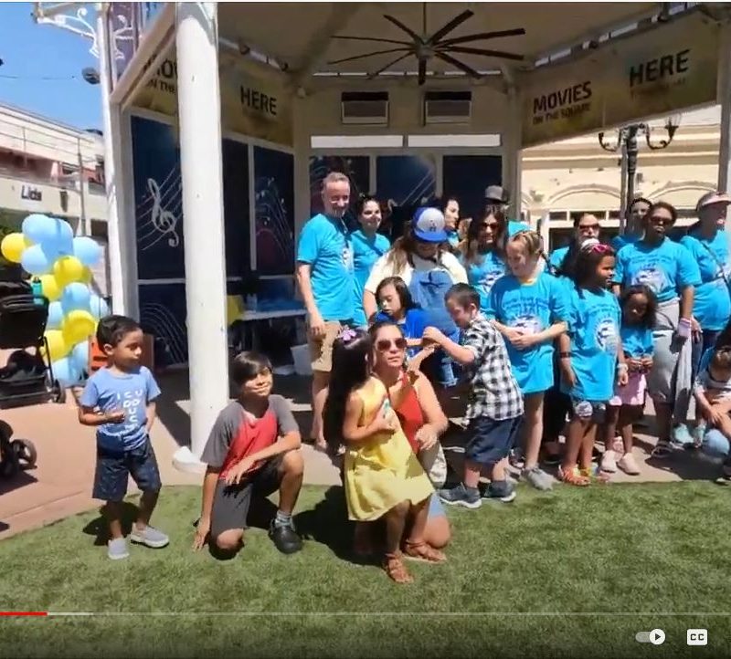 A large group of adults and children in blue shirts stand on a sunny lawn with "HERE" signs overhead.