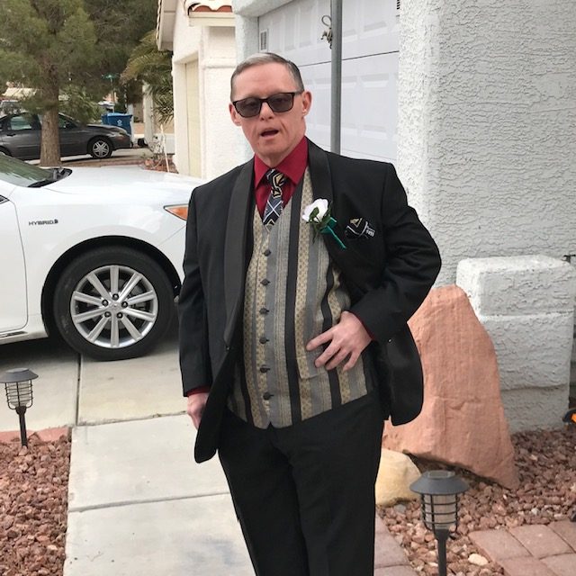 A man in a black tuxedo, red shirt, patterned vest, and sunglasses stands on a sidewalk.