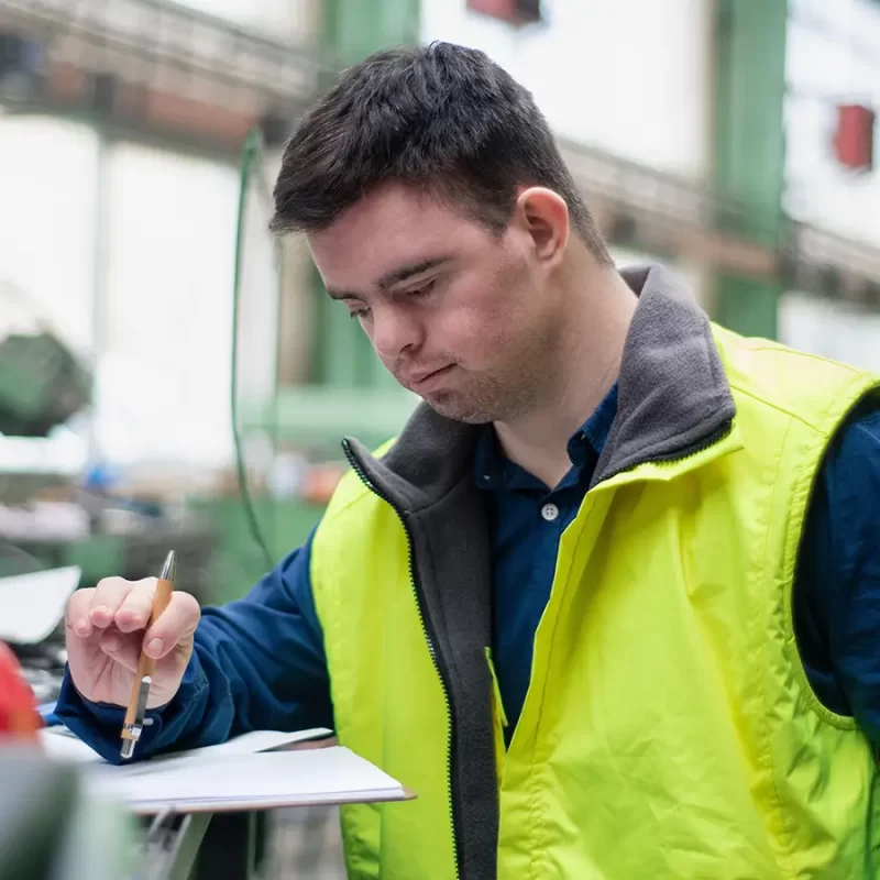A man with Down syndrome in a blue shirt and yellow vest writes on a clipboard in a factory.