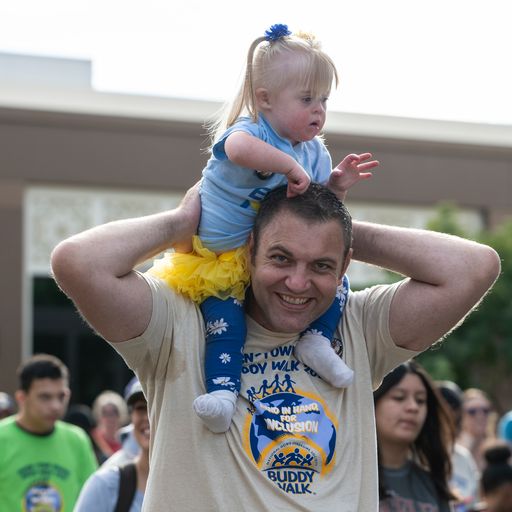 A man with a "Buddy Walk" t-shirt smiles while carrying a blonde toddler with a yellow tutu on his shoulders.