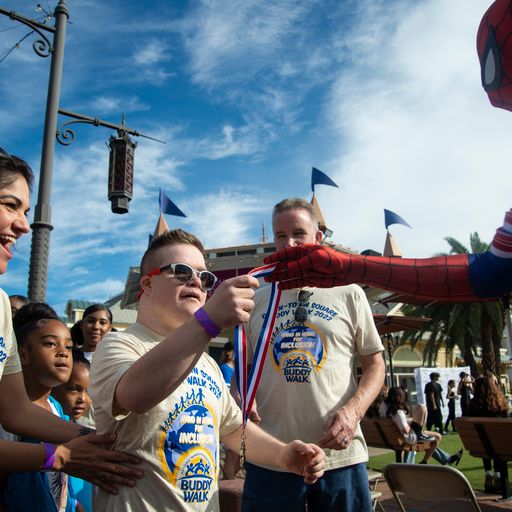 A young man in sunglasses and a "Buddy Walk" shirt receives a medal from a person in a Spiderman costume.