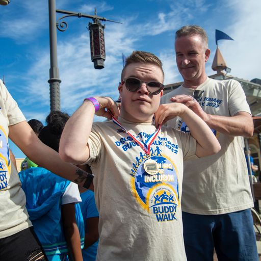 A young man in sunglasses and a "Buddy Walk" t-shirt adjusts a medal, while an older man smiles behind him.