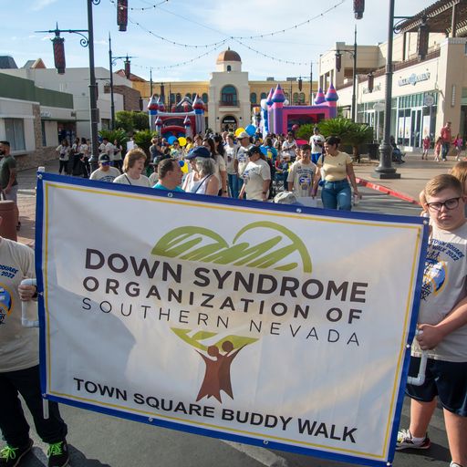 People walk behind a banner that reads "DOWN SYNDROME ORGANIZATION OF SOUTHERN NEVADA TOWN SQUARE BUDDY WALK."