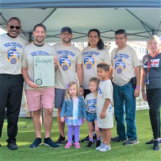Seven adults and three children stand on grass, some wearing "BUDDY WALK 2022" shirts; one man holds a "Proclamation."