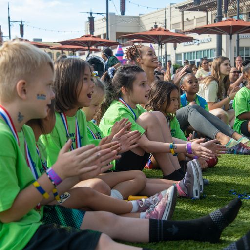 Children and adults in green shirts sit on artificial grass, some clapping, at an outdoor event.