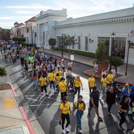 A large group of people, many in yellow shirts, walk down a street past a white building with "H&M" on it.