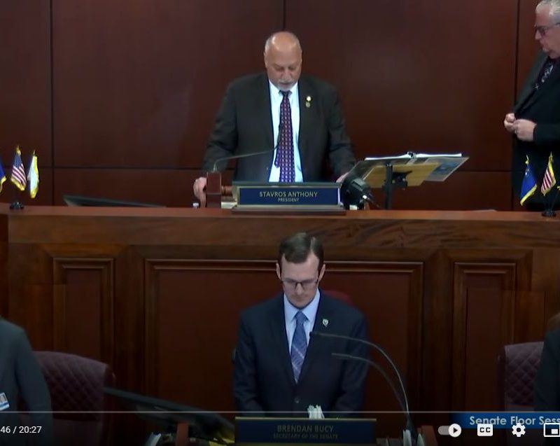 A bald man in a suit stands at a podium labeled "STAVROS ANTHONY PRESIDENT" in a wood-paneled room.