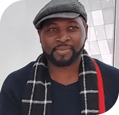 A smiling Black man with a beard wears a gray flat cap, dark sweater, and a black and white plaid scarf.