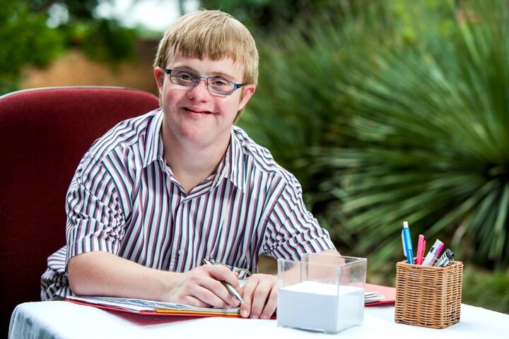 A young man with Down syndrome, wearing glasses and a striped shirt, smiles while writing at a table.