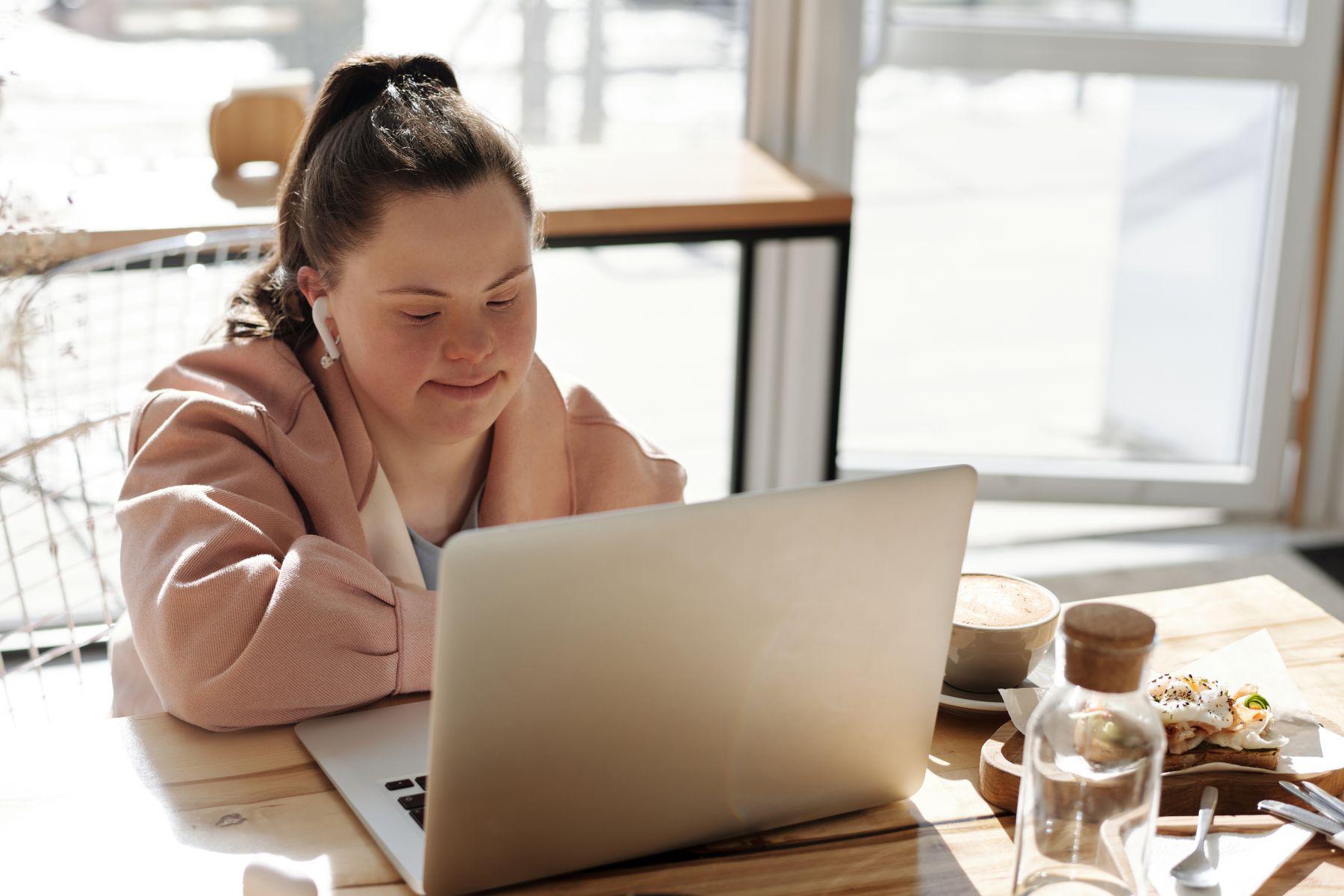 A young woman with Down syndrome, wearing earbuds and a pink jacket, smiles while looking at a laptop.