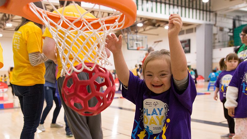 A smiling girl in a purple "PLAY DAY 2019" shirt raises her arms next to a red ball in a basketball hoop.
