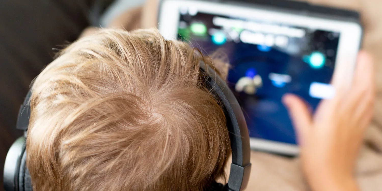A child with light brown hair and black headphones plays a game on a tablet, with their hand on the screen.