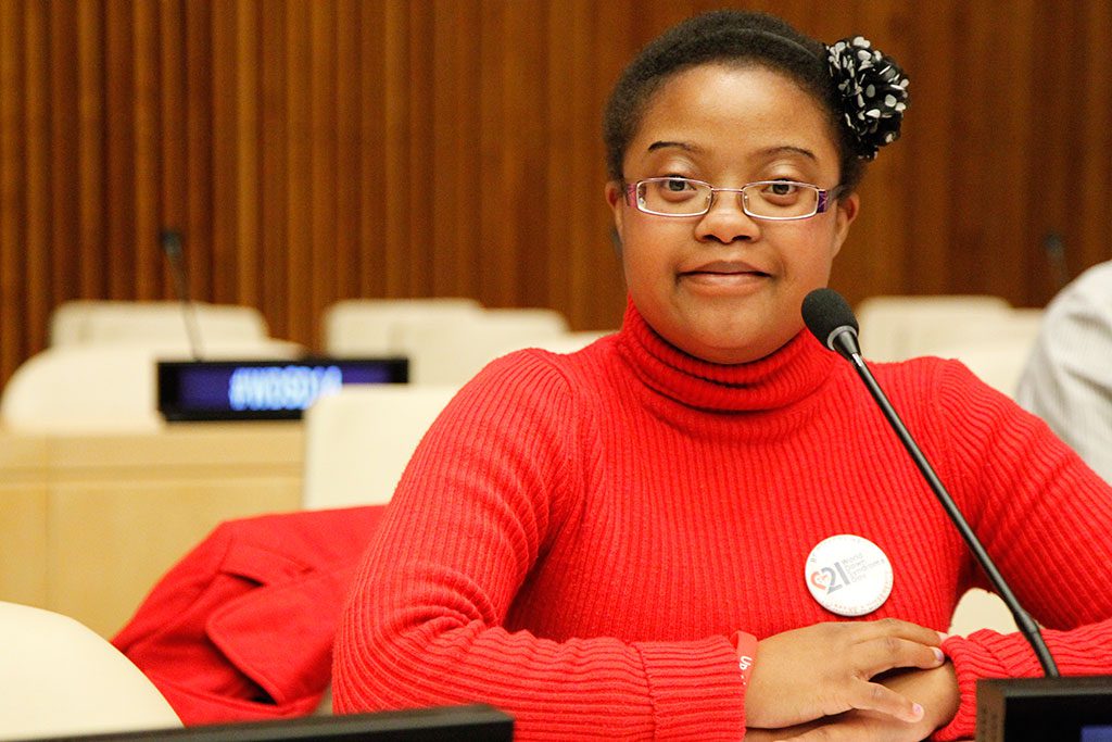 A young woman with glasses and a polka-dot hair accessory wears a red turtleneck and a "21" button, speaking into a microphone.