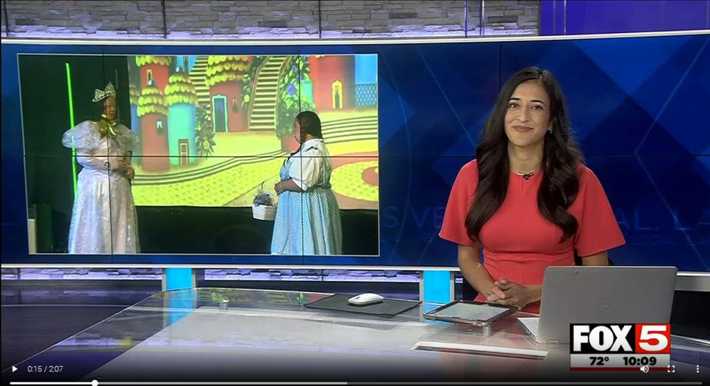 A news anchor in a red dress sits at a desk with a laptop, next to a screen showing two women in costumes.