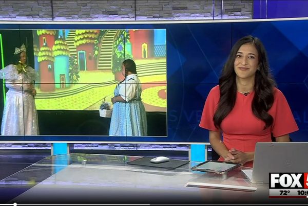 A news anchor in a red dress sits at a desk with a laptop, next to a screen showing two women in costumes.