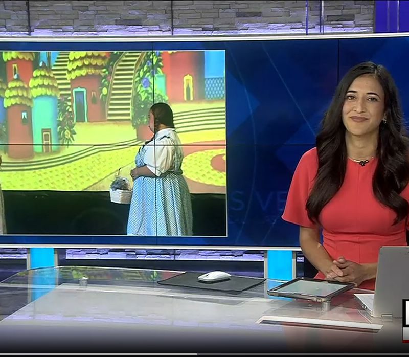A news anchor in a red dress sits at a desk with a laptop, next to a screen showing two women in costumes.