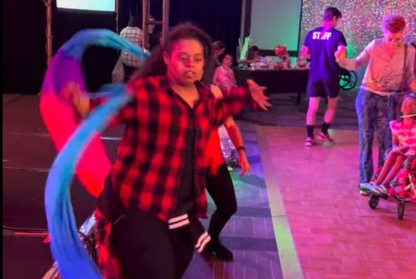 A woman in a red plaid shirt dances with a flowing blue, purple, and red ribbon at a lively event.