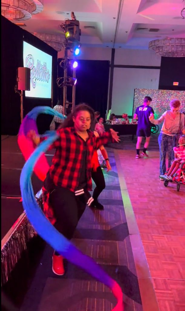 A woman in a red plaid shirt dances with a flowing blue, purple, and red ribbon at a lively event.