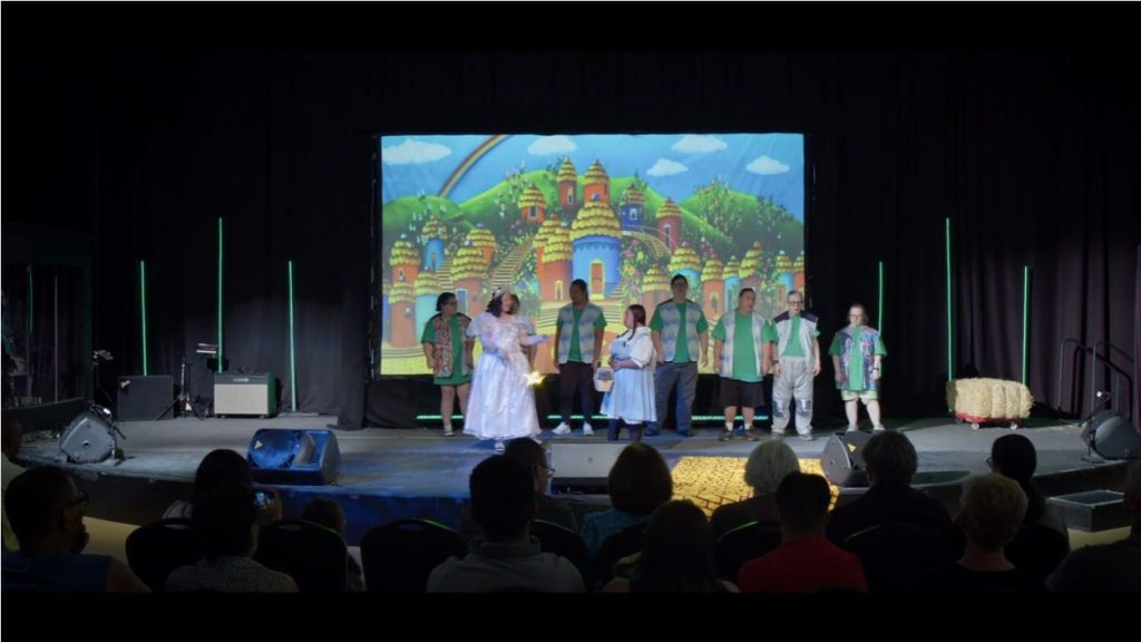 A group of actors in green and white costumes perform on a stage with a colorful cartoon backdrop of a village.