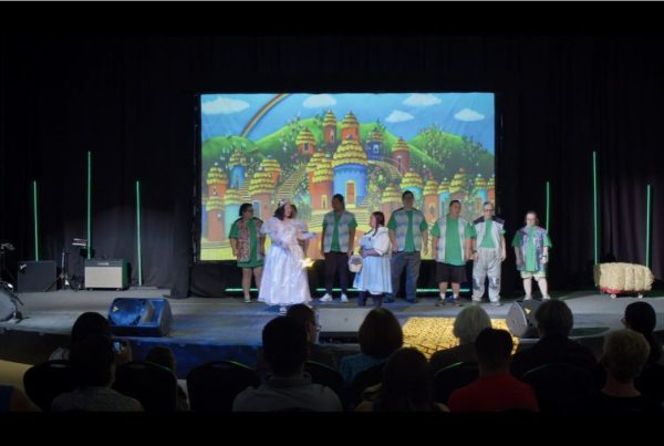 A group of actors in green and white costumes perform on a stage with a colorful cartoon backdrop of a village.