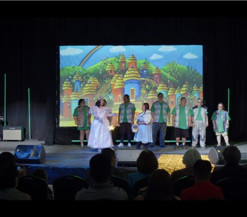A group of actors in green and white costumes perform on a stage with a colorful cartoon backdrop of a village.
