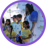 A smiling woman in a crown and blue top stands with five children in yellow shirts, some holding signs.