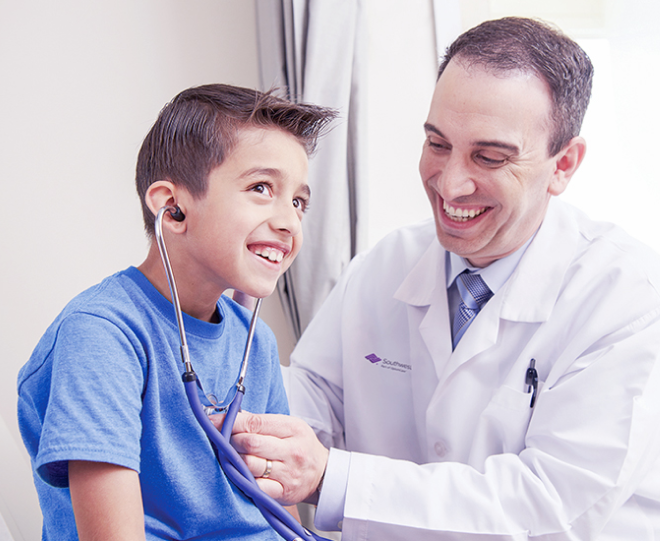 A smiling doctor in a white coat with a "Southwest" logo helps a boy in a blue shirt use a stethoscope.