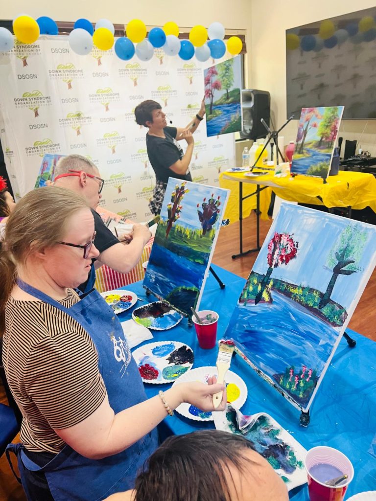 A woman in a blue apron paints a river scene, while an instructor holds up a finished painting.