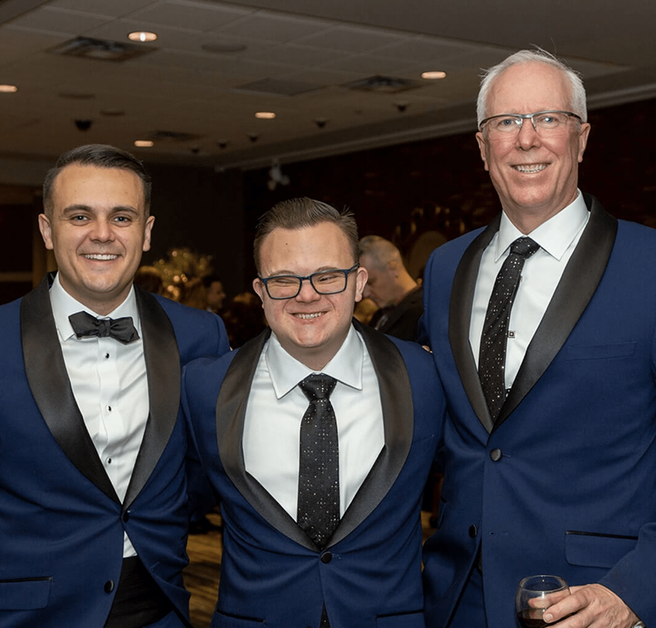 Three smiling men in blue tuxedos with black lapels stand together, one holding a glass of wine.