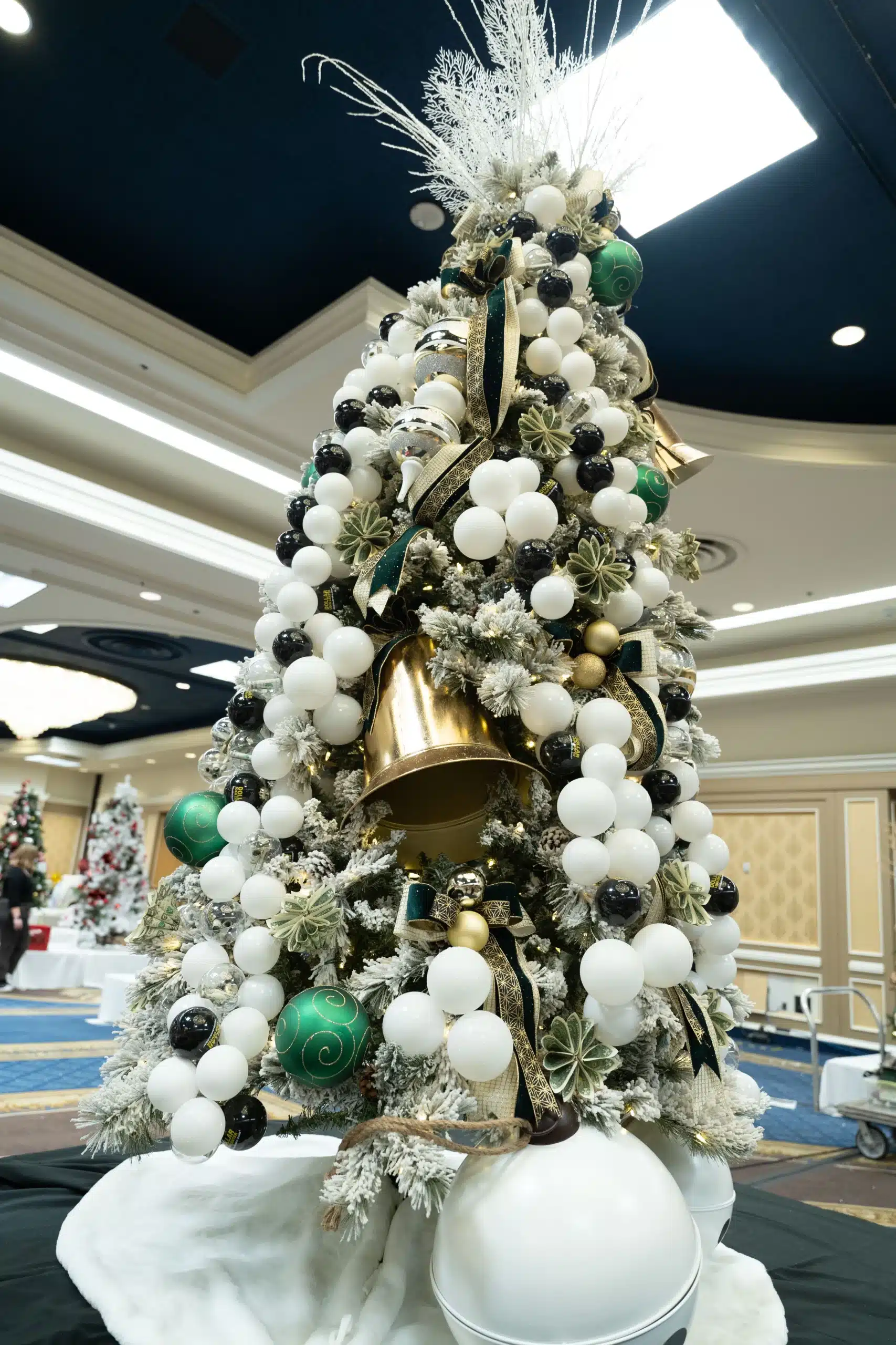 A large, frosted Christmas tree is decorated with white, black, and green ornaments, gold bells, and ribbons.