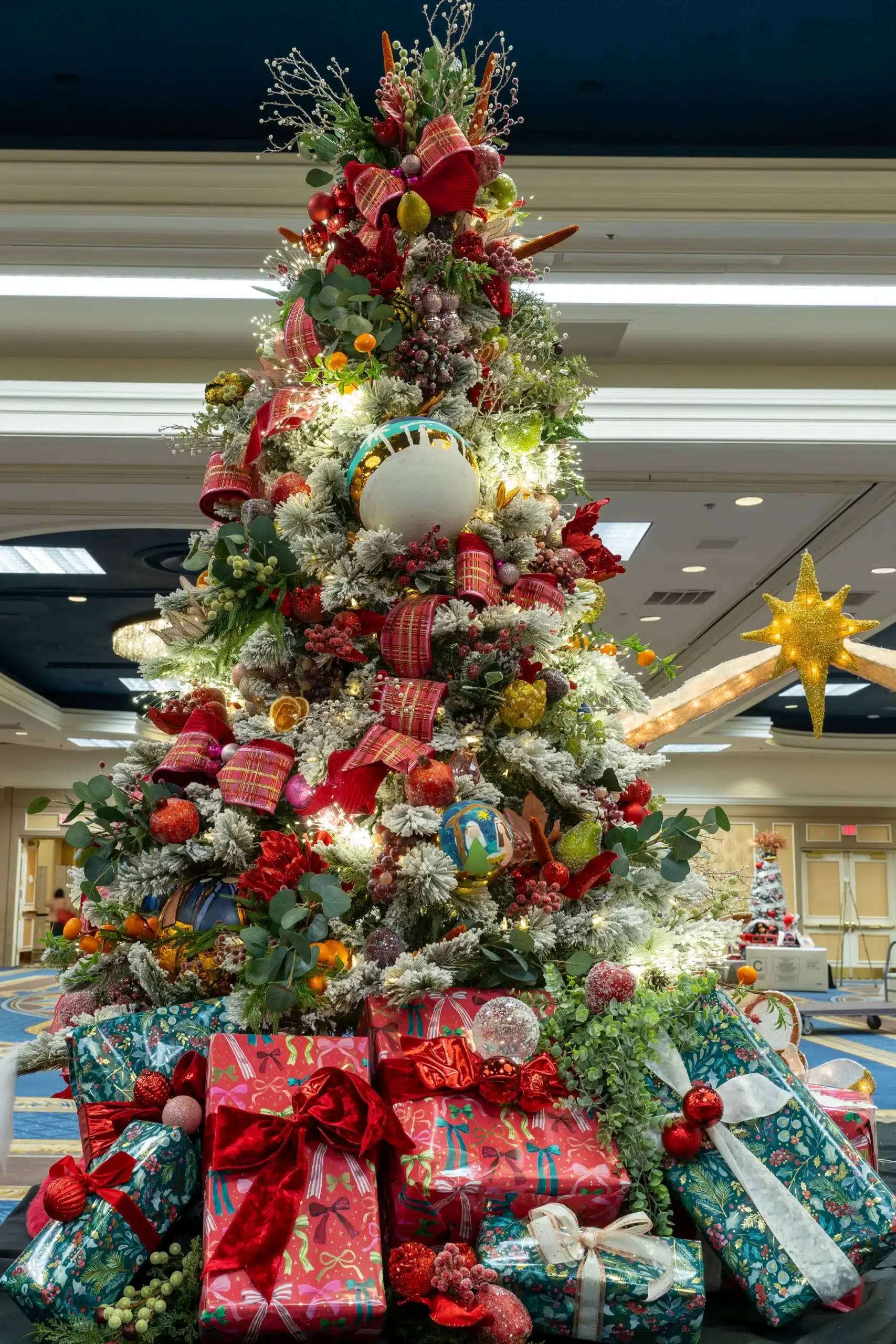 A tall, flocked Christmas tree is decorated with red ribbons, fruit, berries, and lights, surrounded by wrapped gifts.