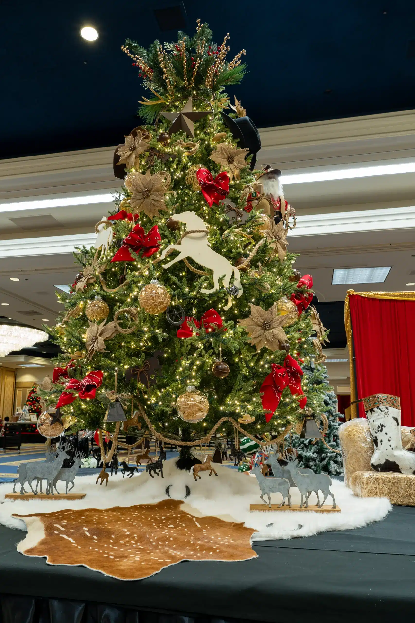 A tall Christmas tree is decorated with white lights, red bows, burlap flowers, and cowboy-themed ornaments.