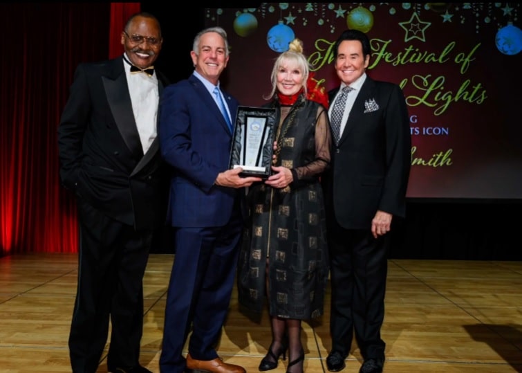 Four people on a stage, two men in suits holding a trophy with a woman, and another man in a tuxedo.