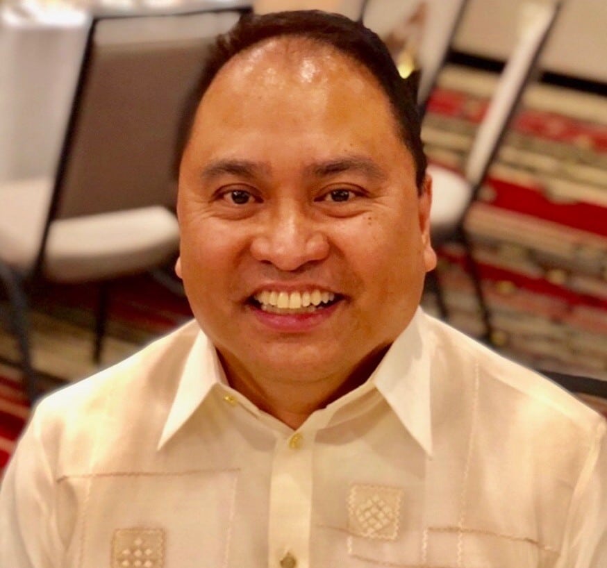 A man with dark hair and a tan complexion smiles broadly, wearing a light-colored, embroidered shirt.