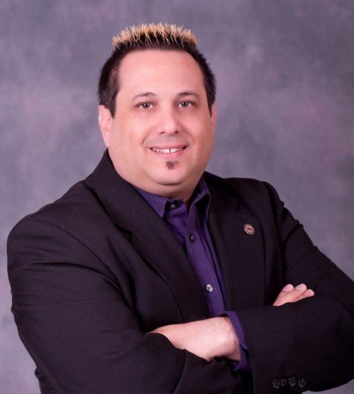 A smiling man with short dark hair and frosted tips wears a purple shirt and black suit jacket.