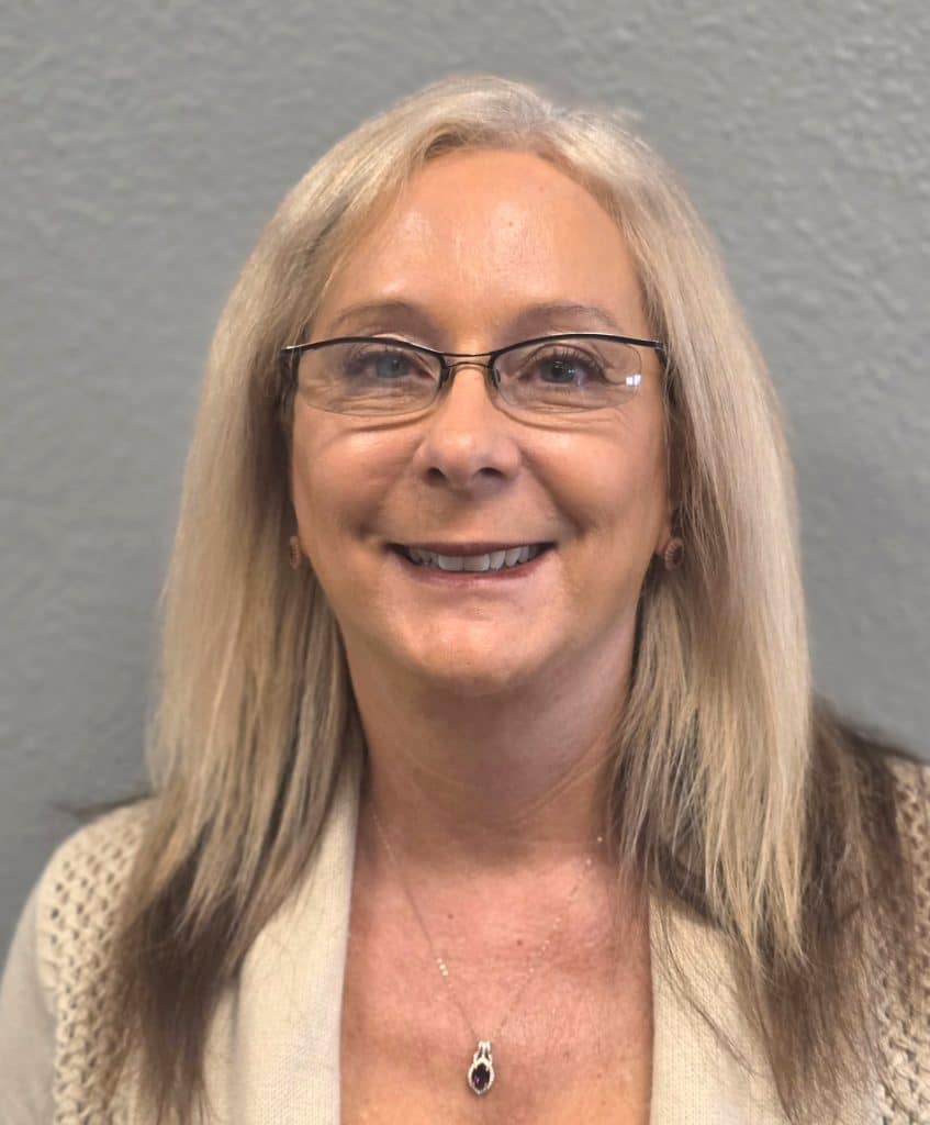 A smiling woman with long blonde hair and glasses wears a beige cardigan and a purple pendant necklace.