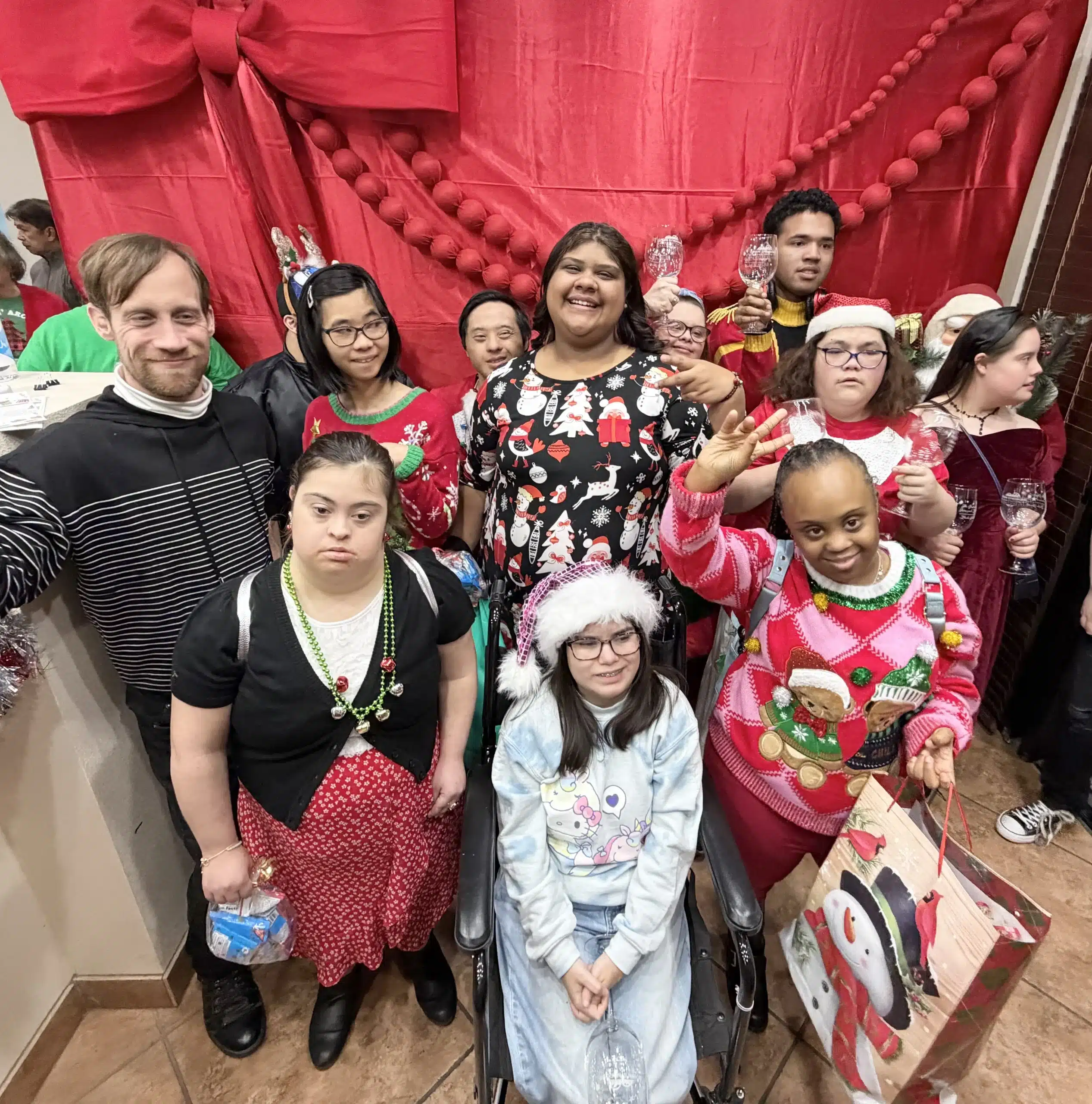 A group of smiling people in festive holiday sweaters and hats pose in front of a giant red bow.