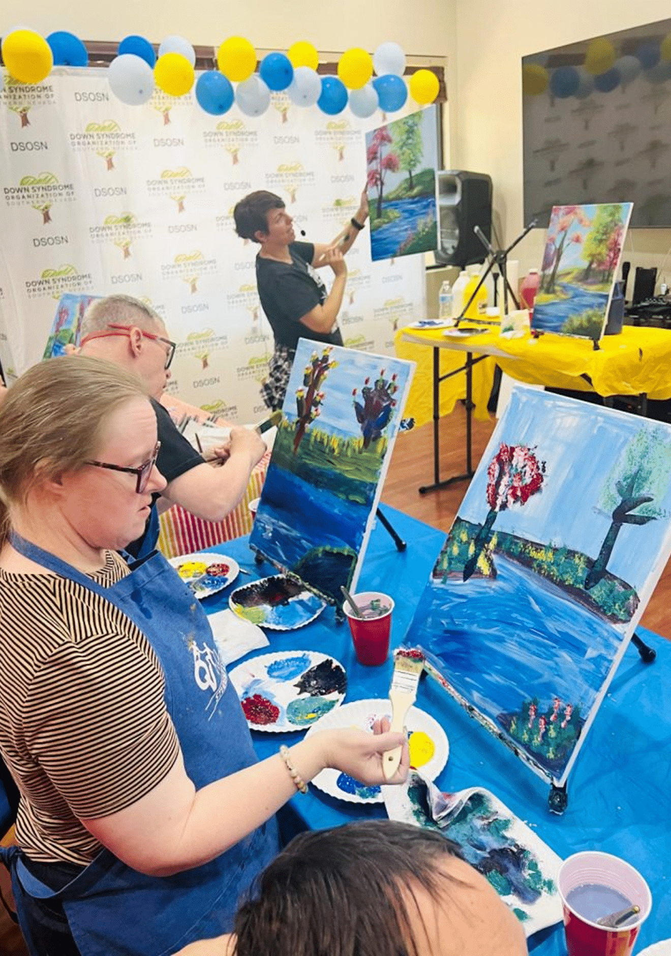 People paint landscapes with trees and rivers on canvases at an event with "DSOSN" banners and balloons.